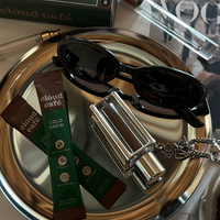 A pair of sunglasses, Cloud Café Collagen cold brew packets, and a silver necklace on a reflective surface.