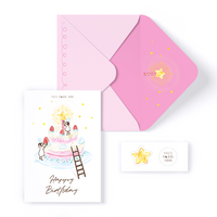 Illustration of 2 fairy-like girls placing cake decorations on a tiered cake, reaching for the lit candle. Greeting Card front message reads "happy birthday". Pastel pink envelope with doodle on the outside, with a hot pink lining and star illustration. Includes a TOJI MOON GOO white and black sticker, with a doodle star sticker.