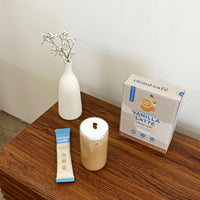 A glass of iced vanilla latte on a wooden counter with a white vase and flowers + a vanilla latte satchet.