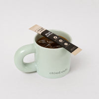 Clöud Café pastel green mug with a rounded thick handle against a white background featuring a Clöud Café Collagen Black Coffee sachet on top