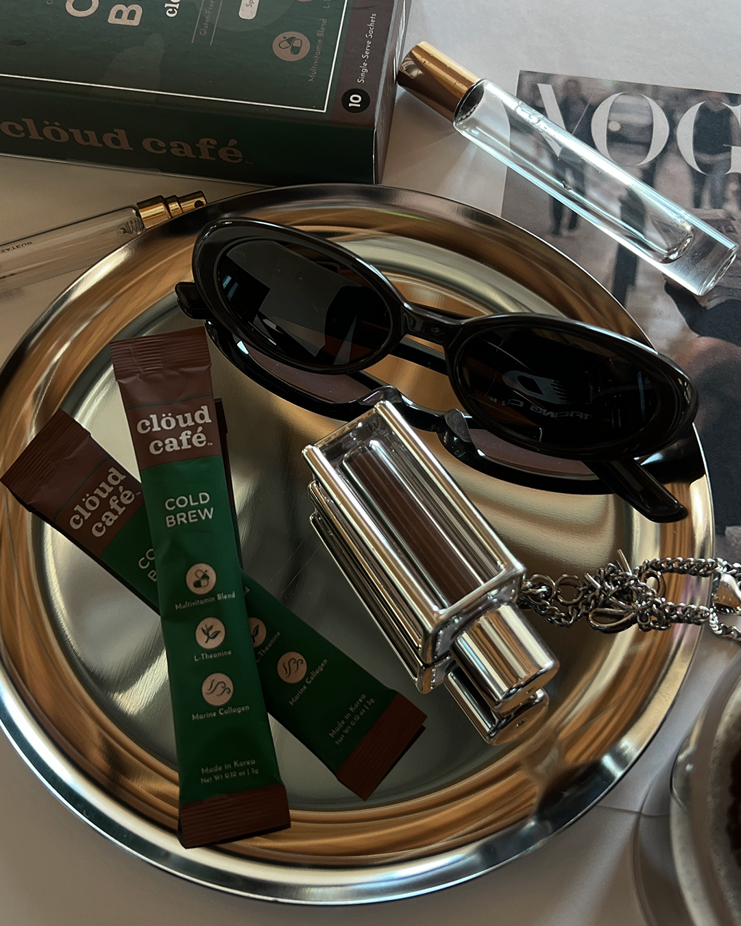 A pair of sunglasses, Cloud Café Collagen cold brew packets, and a silver necklace on a reflective surface.