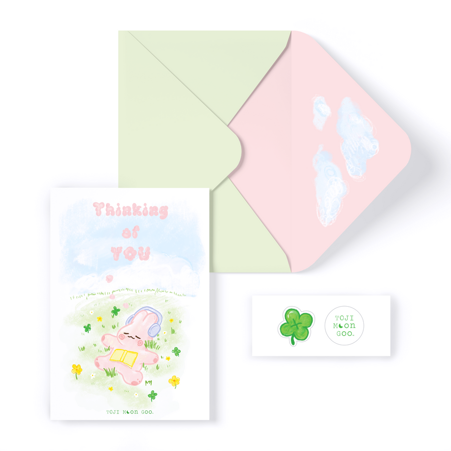 Pink doodle bunny laying on green pastel field. Pink cloud-like "Thinking of YOU" letters in the blue sky. Envelope is light pastel green with a pastel pink interior. Cloud doodles on inside of envelope and card. 1 doodle green clover sticker and 1 TOJI MOON GOO sticker.