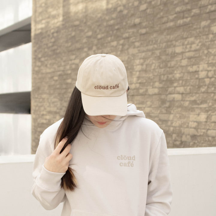 Model wearing beige Clöud Café Merch, including an embroidered baseball cap and beige hoodie.