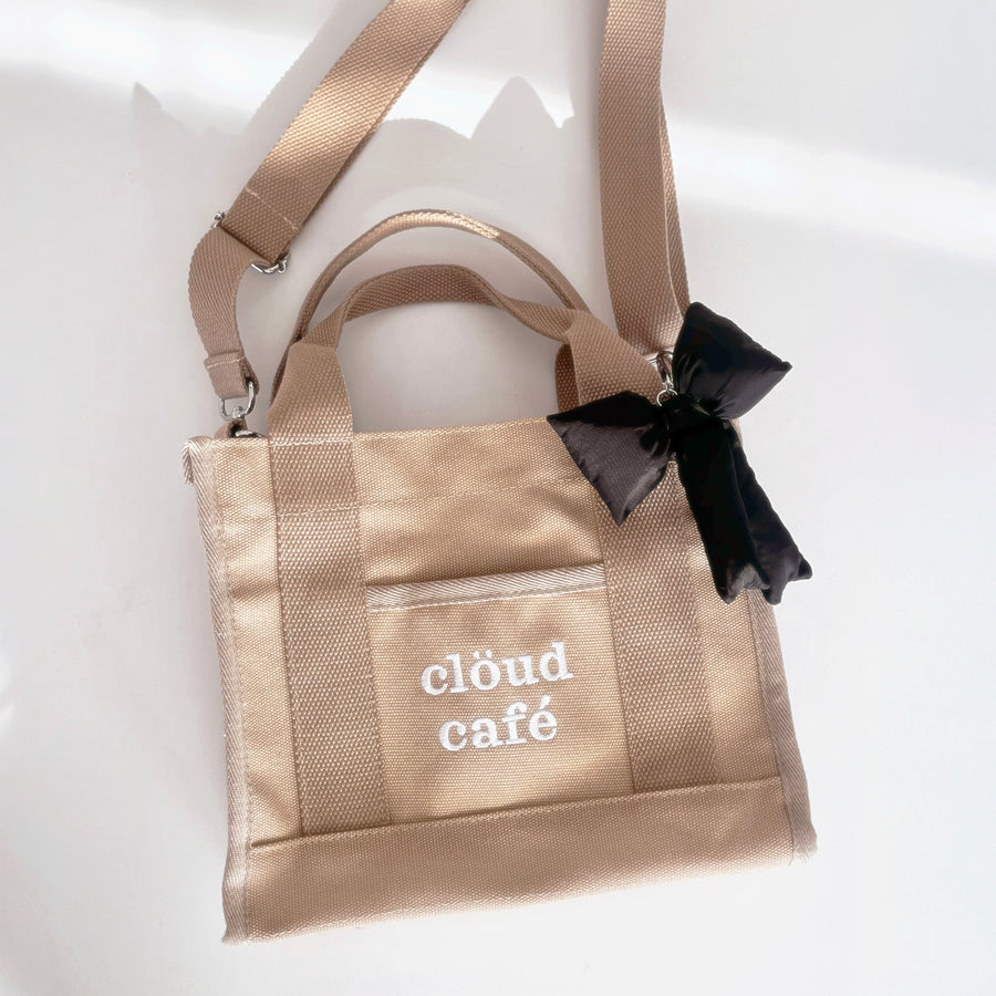 Light tan rectangle canvas tote bag with embroidered "Clöud Café" logo in white. Small pocket in front. Removable long adjustable strap with silver hardware.