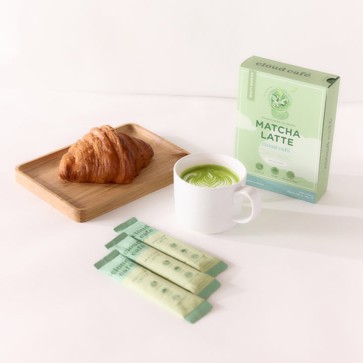 A croissant on a wooden tray, a cup of matcha latte, three matcha latte packets, and a Cloud Cafe matcha latte box arranged on a white surface.