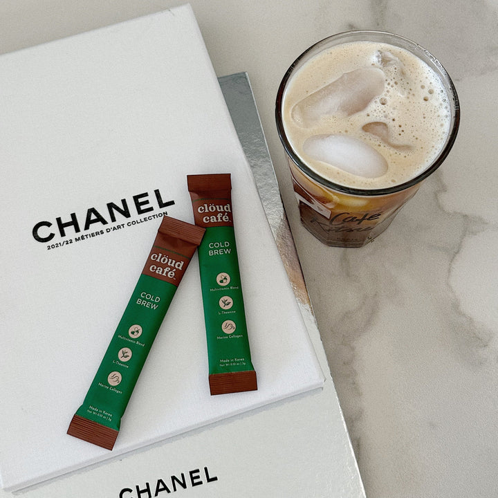 2 clöud café sachets on top of a CHANEL decoration book, next to iced cold brew in a glass cup.