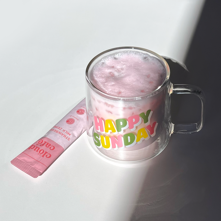 Clear mug with 'Happy Sunday' text filled with pink strawberry milk zero drink, besides strawberry milk zero sachet on white surface