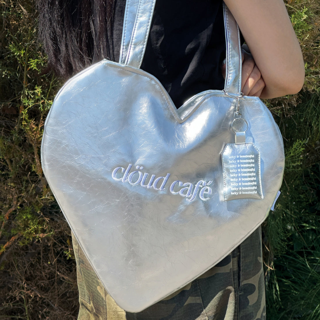 Clöud Café silver heart-shaped logo tote bag displayed in a lifestyle setting surrounded by green foliage